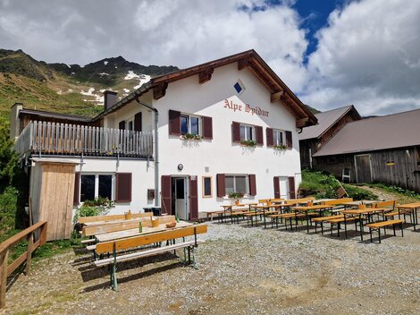 A cozy alpine house named "Alpe Spina". Outside there are many benches and tables in a picturesque mountain landscape.