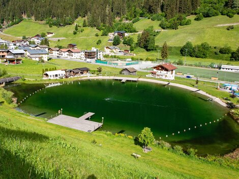 A picturesque pond surrounded by green meadows and mountains. In the background, several houses and facilities are visible.