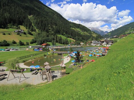 A picturesque park with a swimming lake and green meadows. In the background, mountains and houses can be seen, while people relax and enjoy the sun.