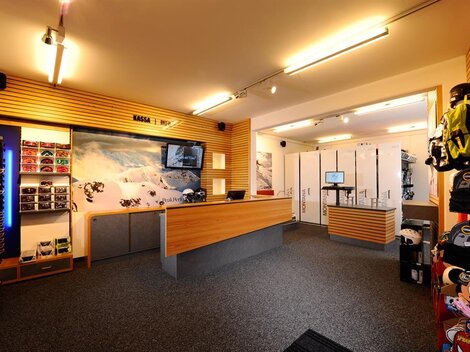 A modern rental space with wooden shelves and a reception desk. In the background, ski equipment and storage areas are visible.
