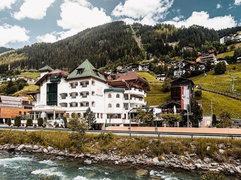 A picturesque hotel on the riverbank, surrounded by green hills and mountains. The buildings are characterized by a lovely rural charm.