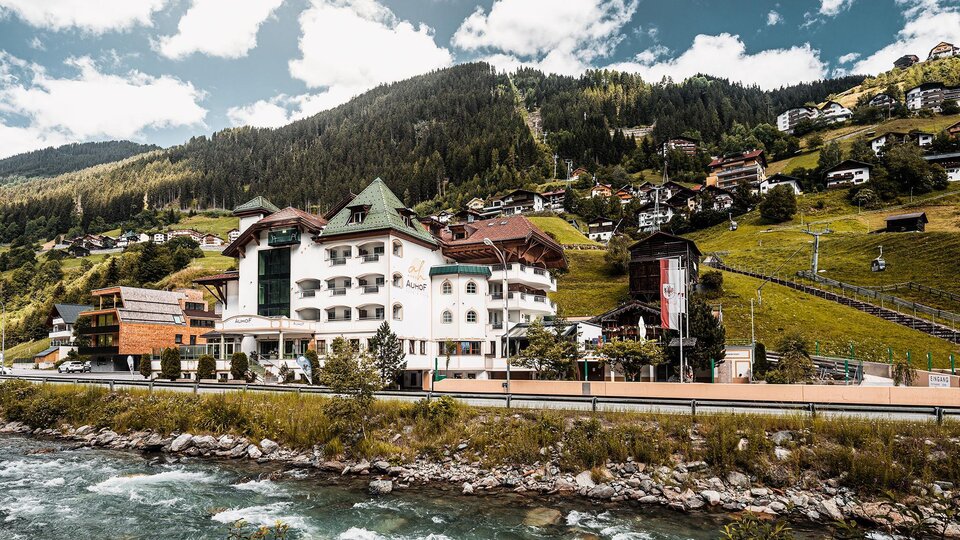 A picturesque hotel on the riverbank, surrounded by green hills and mountains. The buildings are characterized by a lovely rural charm.
