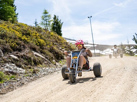 A young person rides down the dusty slope on a tricycle. In the background, there are trees and a clear blue sky.