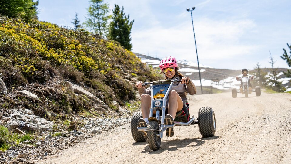 A young person rides down the dusty slope on a tricycle. In the background, there are trees and a clear blue sky.