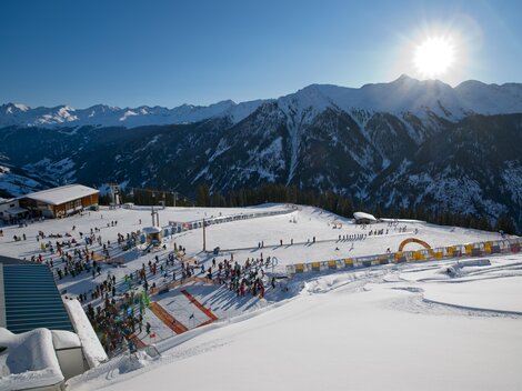 A wintry mountain landscape with skiers and a sunny sky. In the foreground, snow-covered slopes and a busy ski school can be seen.