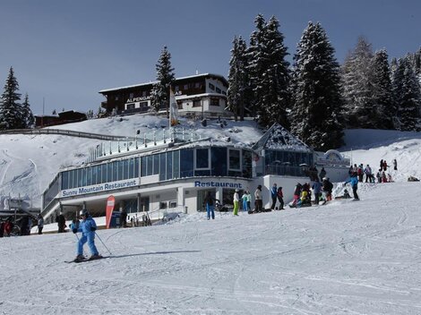 A snow-safe ski course with a modern restaurant in the foreground. Many people are actively enjoying winter fun in a picturesque mountain landscape.