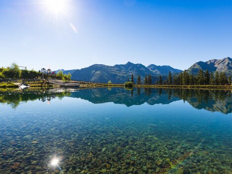 A tranquil lake reflects the mountains and the blue sky. The surroundings are green and sunny.