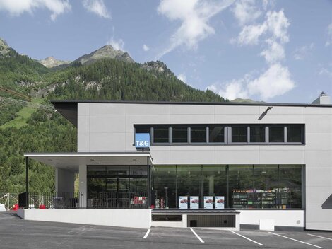 A modern building with large windows and a parking lot in front. In the background, green mountains are visible under a blue sky.