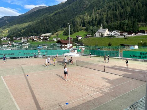 A tennis court with several players and a mountainous landscape in the background. The surroundings are green with typical Alpine architecture.