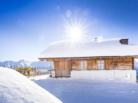 A charming wooden house in the snow with bright sunlight. Snow-covered mountains are visible in the background.