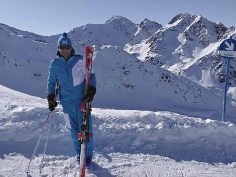 A person stands in the snow with skis in hand. In the background, snow-covered mountains and a clear blue sky can be seen.