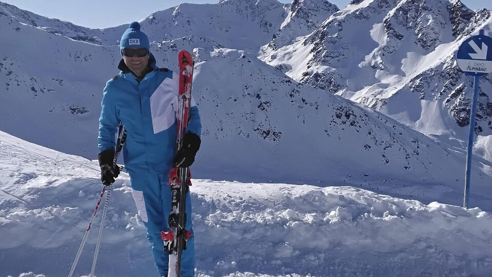 A person stands in the snow with skis in hand. In the background, snow-covered mountains and a clear blue sky can be seen.