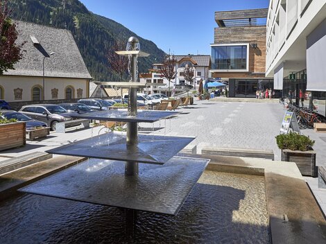 A modern square with a waterfall and seating options. In the background, buildings and parking lots can be seen in a picturesque setting.