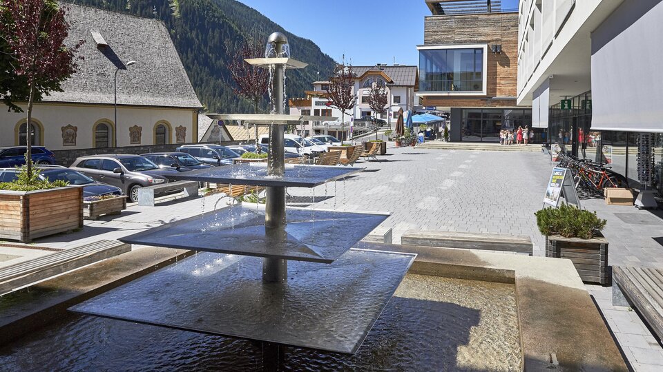 A modern square with a waterfall and seating options. In the background, buildings and parking lots can be seen in a picturesque setting.