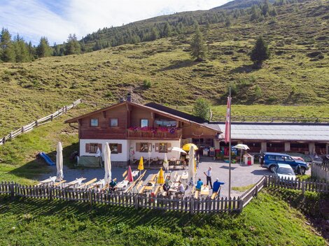 An Alpine inn with a terrace and deck chairs, surrounded by green meadows. In the background, gentle hills and trees rise up.
