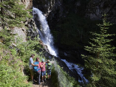 A group of people stands by a waterfall in nature. Surrounded by trees and lush vegetation, they enjoy the view.