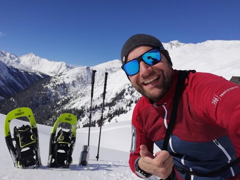 A man takes a selfie in the snowy mountains. In the background, snowy peaks and snowshoes can be seen.