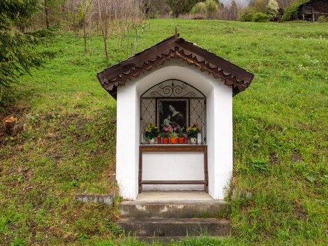 A small chapel on a green hill. In front of the chapel stands a window with flowers and a picture.