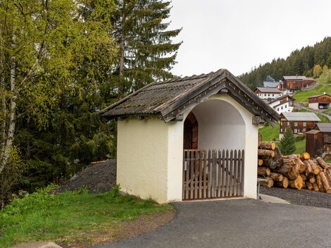 A small, charming cottage with an angled roof stands by a path. In the background, there are wood piles and some houses in a wooded mountain landscape. | © Bernhard Gruber – Photography