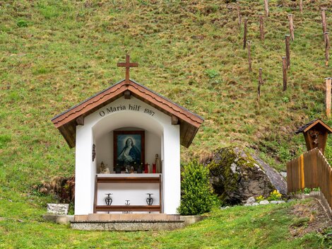 A small chapel with a cross and a picture of the Virgin Mary. It stands in the middle of a green meadow.
