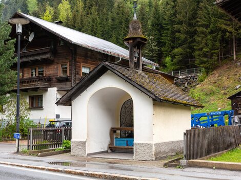 A small white chapel with a bell tower stands by the road. In the background, typical alpine wooden houses and trees can be seen. | © Bernhard Gruber – Photography